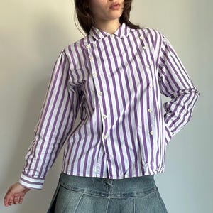 May include: A long-sleeved button-down shirt with vertical purple and white stripes. The shirt has a double-breasted design with multiple buttons and a rounded collar. The cuffs and collar are white.