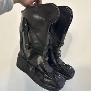 Puede incluir: Un par de botas negras de media caña con parte superior sintética lisa, cierre frontal de cordones con ojales metálicos plateados y suela de goma texturizada con patrón. Las botas tienen una caña alta estructurada y una lengüeta de tela gris oscuro.