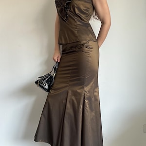 May include: A two-piece brown satin outfit. The top is sleeveless with a ruffled neckline. The skirt is a midi length with a flared hem. A small black handbag and black heels complete the look. The outfit is a formal wear ensemble.