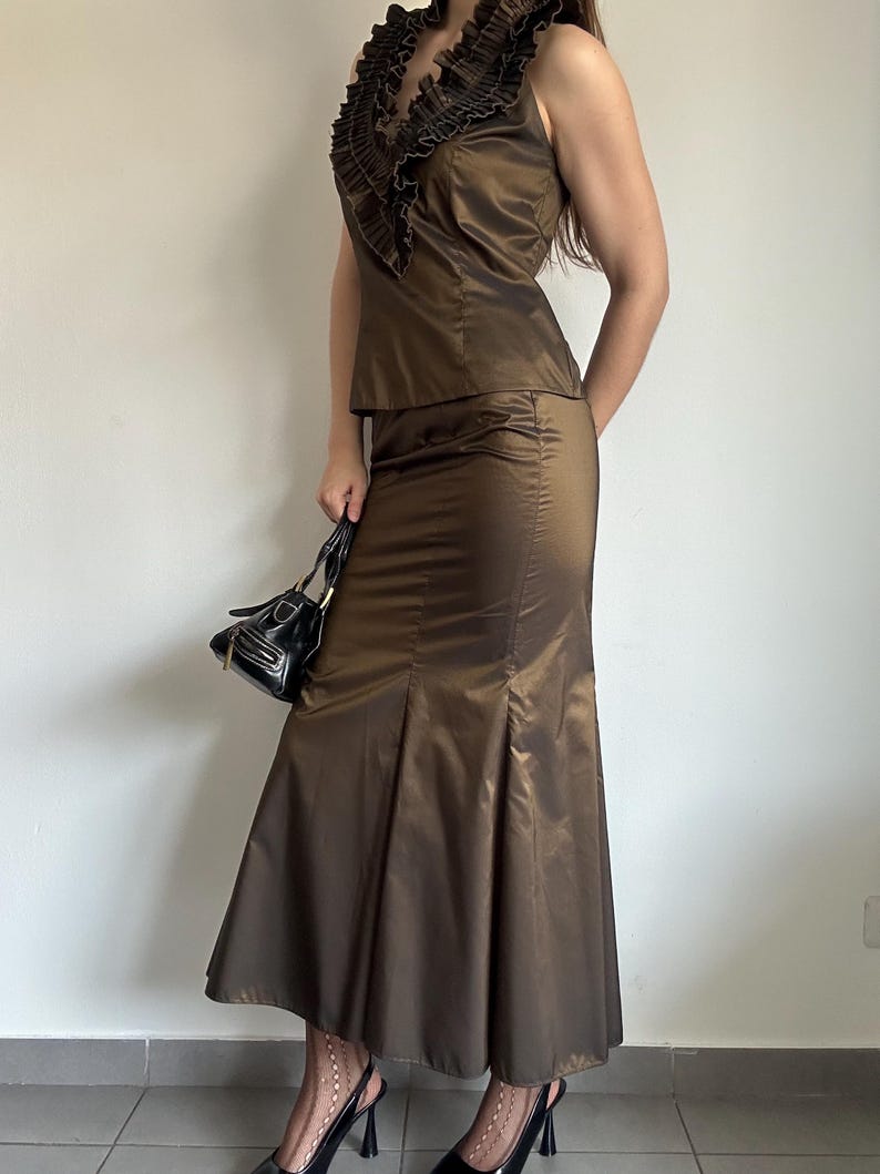 May include: A two-piece brown satin outfit. The top is sleeveless with a ruffled neckline. The skirt is a midi length with a flared hem. A small black handbag and black heels complete the look. The outfit is a formal wear ensemble.