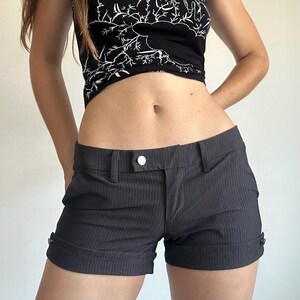 May include: Dark gray pinstriped shorts featuring a low-rise waist, a single silver-toned button closure, belt loops, and cuffed hems secured with small buttons on the outer sides. The fabric has a subtle vertical pinstripe pattern throughout.