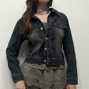Könnte beinhalten: Dunkelblaue Jeansjacke mit Knopfleiste, zwei Brusttaschen und langen Ärmeln. Die Jacke hat einen verwaschenen, abgenutzten Look. Die Jacke wird mit einem grauen Oberteil und Rock kombiniert. Eine silberfarbene Halskette ist ebenfalls sichtbar.