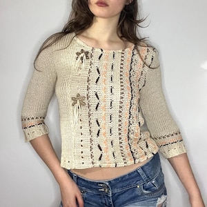 May include: Beige knit top with three-quarter sleeves. The top features a round neckline and is adorned with brown, orange, and black patterned details. Two decorative flower accents with fringe are on the front. The cuffs have a decorative trim.
