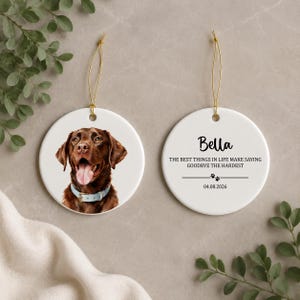 Peut inclure: Deux ornements ronds en céramique blanche avec des cordons dorés. L'un présente un chien Labrador marron portant un collier bleu clair. L'autre affiche le texte Bella, The best things in life make saying goodbye the hardest, et 04.08.2026.