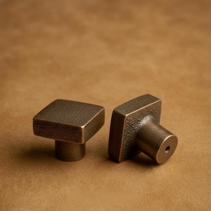 May include: Two bronze-coloured square cabinet knobs with a textured finish. The knobs have a simple, modern design, with a square face and a cylindrical base. The knobs are likely made of metal and are suitable for furniture or cabinetry.