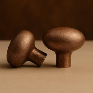 Peut inclure: Deux boutons de meuble en bronze avec une texture martelée. Les boutons ont une forme ovale arrondie avec une courte tige cylindrique. Les boutons sont présentés sur un fond neutre, mettant en valeur leur finition métallique.