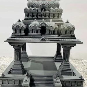 Può includere: Un dettagliato tempio in miniatura grigio con più cupole e pilastri. La parte superiore del tempio presenta accenti dorati. La struttura comprende una serie di scale che conducono a una piattaforma aperta. Il design generale è intricato e simmetrico.