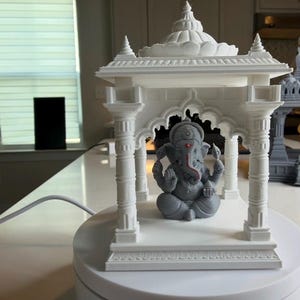 Può includere: Una statua grigia del dio indù Ganesha, seduta all'interno di una struttura di tempio bianca e ornata. La statua ha un segno rosso sulla fronte e una proboscide rossa. Il tempio presenta colonne e un tetto dettagliato, creando un tema religioso o spirituale.