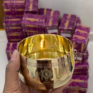Puede incluir: Un cuenco de metal dorado pulido de forma cilíndrica, con líneas horizontales grabadas y un emblema circular central. El cuenco tiene una pequeña anilla de metal en el lateral y una superficie lisa y reflectante que capta la luz.