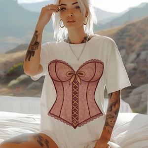 May include: White t-shirt featuring a pink corset design with a bow and lace-up detail. The corset graphic has a scalloped edge and is centered on the shirt. The shirt is a casual, oversized style.