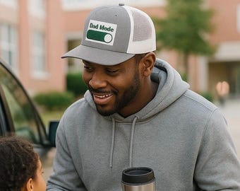 May include: A grey trucker cap with a white mesh back and a green and white patch that reads "Dad Mode". The hat is worn by a person wearing a grey hoodie and holding a stainless steel travel mug.