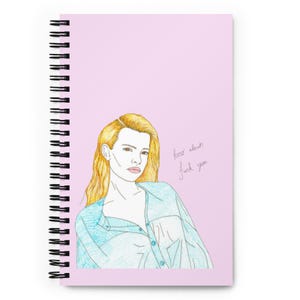May include: A spiral-bound notebook with a light pink cover featuring a colored pencil illustration of a person with blonde hair wearing a light blue button-down shirt. The text "how about fuck you" is written in cursive next to the figure.