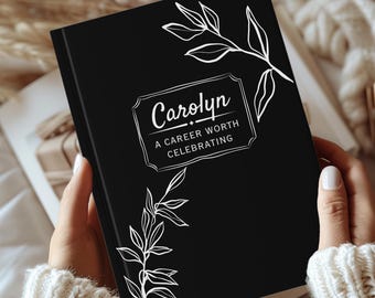 Puede incluir: Un diario negro con detalles florales blancos y el nombre "Carolyn" en un marco decorativo. El texto debajo dice "A Career Worth Celebrating". El diario se sostiene abierto, revelando las páginas interiores.