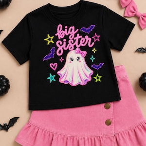 May include: A black short-sleeve t-shirt featuring the text big sister in pink glittery script. The design includes a white ghost with a pink bow, purple bats, and colorful stars and hearts. Paired with a pink skirt featuring two buttons.