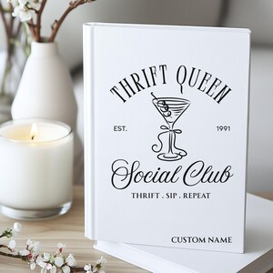 K&ouml;nnte beinhalten: Ein wei&szlig;es Notizbuch mit dem Text "Thrift Queen Social Club" in schwarzer Schrift. Das Notizbuch zeigt eine Martini-Glas-Illustration und die Worte "Thrift. Sip. Repeat." Eine brennende Kerze und Blumen sind im Hintergrund.