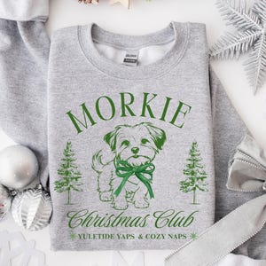May include: A gray sweatshirt with a green graphic of a dog wearing a bow, the word "Morkie", and the text "Christmas Club". The sweatshirt also features two Christmas trees and the phrase "Yuletide Yaps & Cozy Naps".