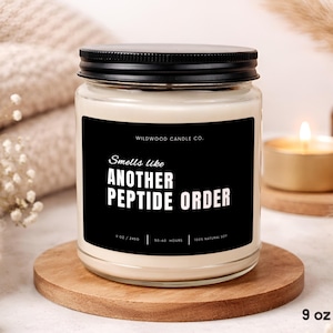 May include: A clear glass candle jar with a black lid and a black label featuring the white text "WILDWOOD CANDLE CO." and "Smells like ANOTHER PEPTIDE ORDER". The label also displays "9 OZ / 245G", "50-60 HOURS", and "100% NATURAL SOY" in white text.