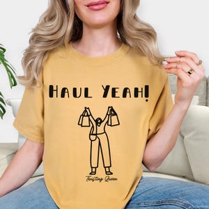May include: Mustard yellow t-shirt with the text "HAUL YEAH!" in black block letters. Below the text is a black line drawing of a person holding shopping bags. The words "Thrifting Queen" are printed below the drawing.