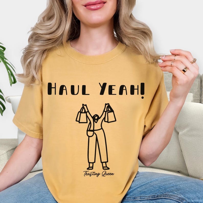 May include: Mustard yellow t-shirt with the text "HAUL YEAH!" in black block letters. Below the text is a black line drawing of a person holding shopping bags. The words "Thrifting Queen" are printed below the drawing.