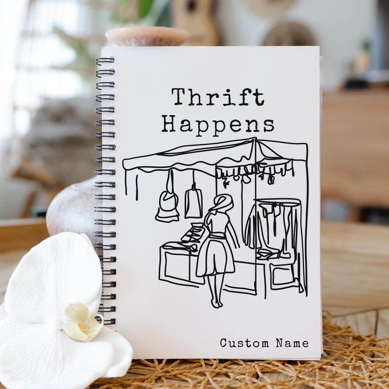K&ouml;nnte beinhalten: Ein wei&szlig;es Spiralnotizbuch mit dem Text "Thrift Happens" &uuml;ber einer Schwarz-Wei&szlig;-Illustration einer Marktszene. Die Worte "Custom Name" stehen unten. Das Notizbuch liegt auf einer Holzoberfl&auml;che mit einer wei&szlig;en Blume und einem beigen Stein.