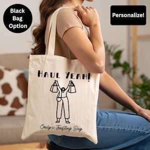 May include: Cream-colored tote bag with the text "HAUL YEAH!" and a graphic of a person holding shopping bags. The bag also has the text "Emily's Thrifting Bag". The bag has long handles and is being held by a person.