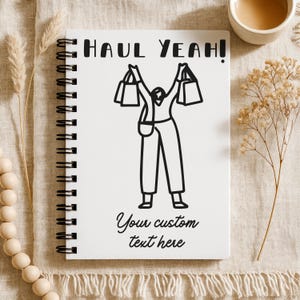 K&ouml;nnte beinhalten: Ein wei&szlig;es Spiralnotizbuch mit den Worten "HAUL YEAH!" und "Your custom text here" auf dem Einband. Der Einband zeigt eine Schwarz-Wei&szlig;-Illustration einer Person, die Einkaufstaschen h&auml;lt. Eine Tasse Kaffee und dekorative Elemente befinden sich im Hintergrund.