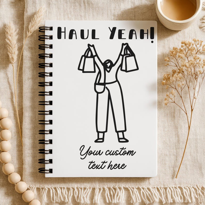 K&ouml;nnte beinhalten: Ein wei&szlig;es Spiralnotizbuch mit den Worten "HAUL YEAH!" und "Your custom text here" auf dem Einband. Der Einband zeigt eine Schwarz-Wei&szlig;-Illustration einer Person, die Einkaufstaschen h&auml;lt. Eine Tasse Kaffee und dekorative Elemente befinden sich im Hintergrund.