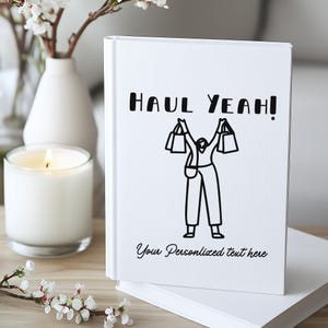 K&ouml;nnte beinhalten: Ein wei&szlig;es Notizbuch mit dem Text "HAUL YEAH!" und einer Grafik einer Person, die Einkaufstaschen h&auml;lt. Der Satz "Your Personalized text here" steht unter der Grafik. Eine brennende Kerze, eine Vase und Blumen sind im Hintergrund.