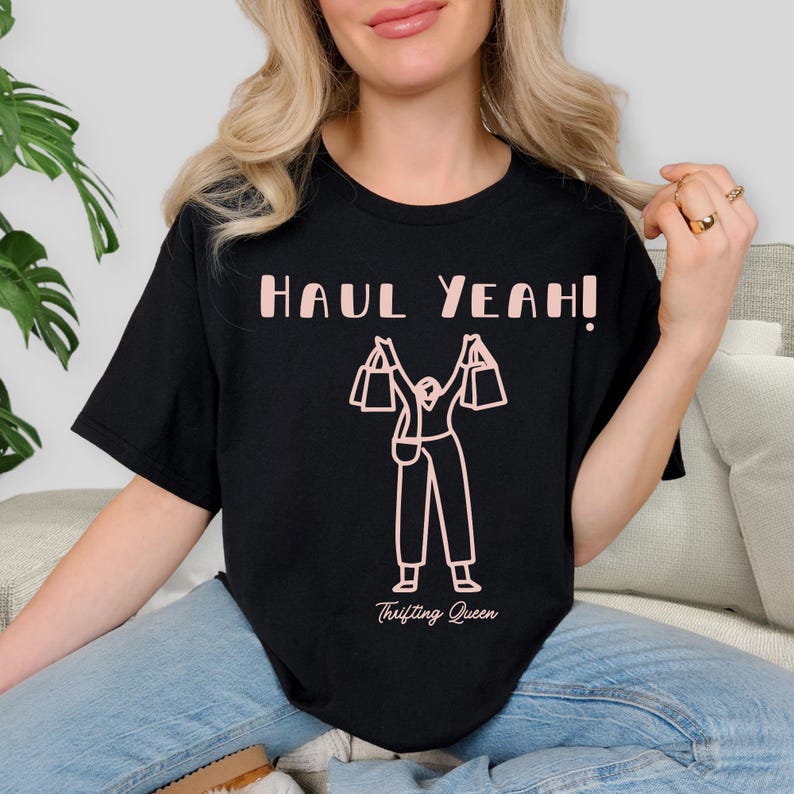 May include: Black t-shirt with the text "HAUL YEAH!" in pink, above a pink graphic of a person holding shopping bags. The words "Thrifting Queen" are printed below the graphic.