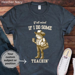 May include: Navy blue t-shirt featuring a cartoon frog in a cowboy hat, carrying a coffee cup and a bag marked "TEACHER." The text says "Y'all mind IF I DO SOME TEACHIN'."