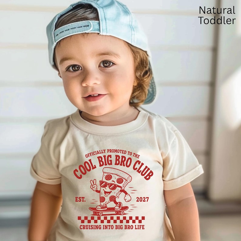 May include: A cream-colored t-shirt featuring a red graphic of a pizza slice wearing sunglasses on a skateboard. The text reads: OFFICIALLY PROMOTED TO THE COOL BIG BRO CLUB EST. 2027 CRUISING INTO BIG BRO LIFE. A light blue baseball cap is worn.