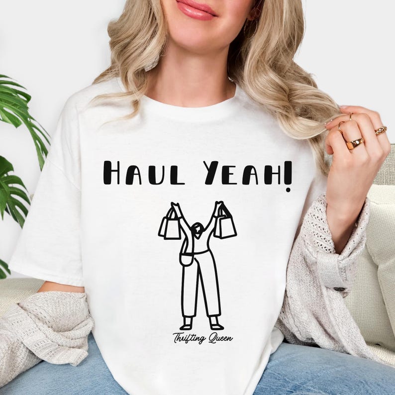 May include: White t-shirt with the text "HAUL YEAH!" in black, above a black and white illustration of a person holding shopping bags. The words "Thrifting Queen" are below the illustration.