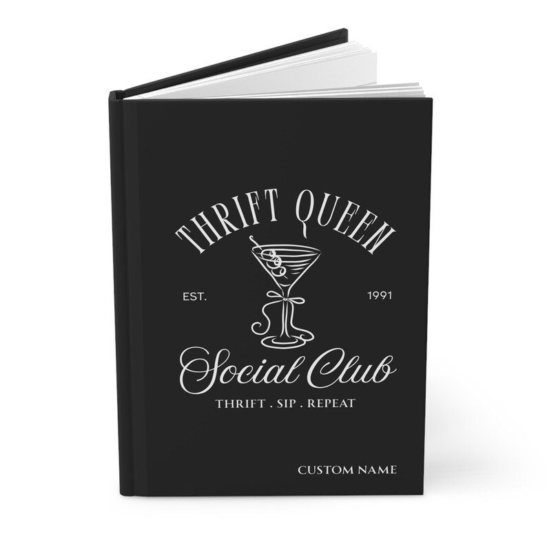 K&ouml;nnte beinhalten: Schwarzes Notizbuch mit dem wei&szlig;en Schriftzug "THRIFT QUEEN Social Club". Das Design beinhaltet eine Cocktailglas-Illustration und die Worte "THRIFT. SIP. REPEAT." Das Notizbuch ist ge&ouml;ffnet und zeigt wei&szlig;e Seiten.