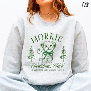 May include: Ash-colored sweatshirt with a green graphic featuring the word "Morkie" above a cartoon dog wearing a bow. Below the dog is "Christmas Club" and "Yuletide Yaps & Cozy Naps". Two green Christmas trees flank the dog.