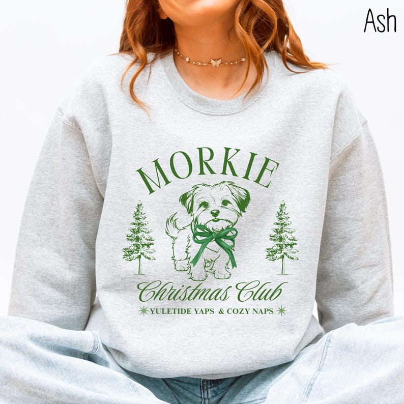 May include: Ash-colored sweatshirt with a green graphic featuring the word "Morkie" above a cartoon dog wearing a bow. Below the dog is "Christmas Club" and "Yuletide Yaps & Cozy Naps". Two green Christmas trees flank the dog.