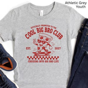 May include: An athletic grey short-sleeve t-shirt featuring a red graphic of a pizza slice on a skateboard. The text reads: OFFICIALLY PROMOTED TO THE COOL BIG BRO CLUB, EST. 2027, and CRUISING INTO BIG BRO LIFE. The pizza slice wears sunglasses.