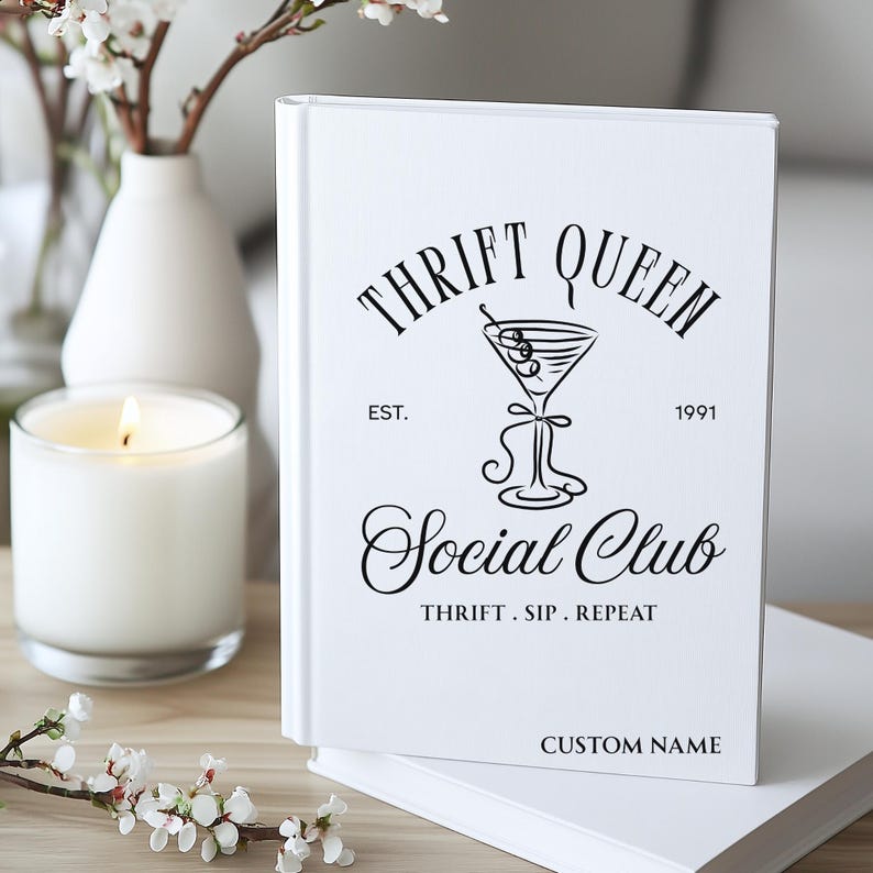 K&ouml;nnte beinhalten: Ein wei&szlig;es Notizbuch mit dem Text "Thrift Queen Social Club" in schwarzer Schrift. Das Notizbuch zeigt eine Martini-Glas-Illustration und die Worte "Thrift. Sip. Repeat." Eine brennende Kerze und Blumen sind im Hintergrund.