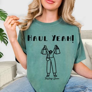 May include: A seafoam green t-shirt with the words "HAUL YEAH!" in bold black letters. Below the text is a black graphic of a person holding shopping bags. The words "Thrifting Queen" are printed below the graphic.