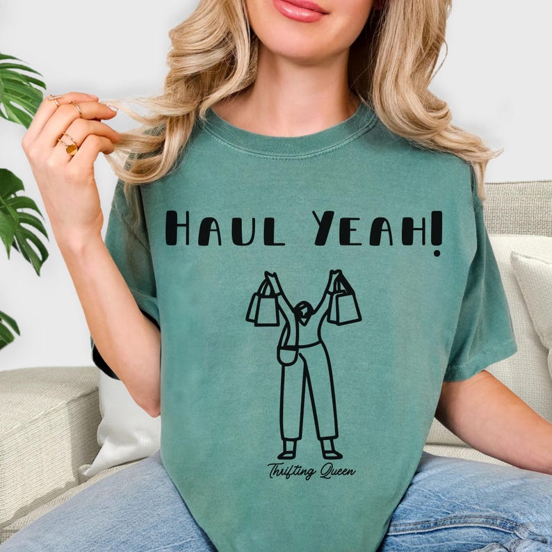 May include: A seafoam green t-shirt with the words "HAUL YEAH!" in bold black letters. Below the text is a black graphic of a person holding shopping bags. The words "Thrifting Queen" are printed below the graphic.
