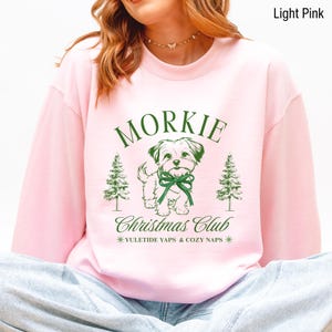 May include: Light pink sweatshirt with a green graphic of a Morkie dog wearing a bow, flanked by Christmas trees. The text "Morkie Christmas Club" and "Yuletide Yaps & Cozy Naps" are also in green.