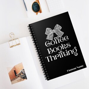 K&ouml;nnte beinhalten: Ein schwarzes Spiralnotizbuch mit den Worten "Coffee Books Thrifting" in wei&szlig;er Schrift, gekr&ouml;nt von einer Schleifengrafik. Die Worte "Custom Name" stehen unten. Das Notizbuch ist auf einer wei&szlig;en Oberfl&auml;che ge&ouml;ffnet.