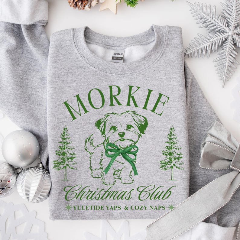 May include: A gray sweatshirt with a green graphic of a dog wearing a bow, the word "Morkie", and the text "Christmas Club". The sweatshirt also features two Christmas trees and the phrase "Yuletide Yaps & Cozy Naps".