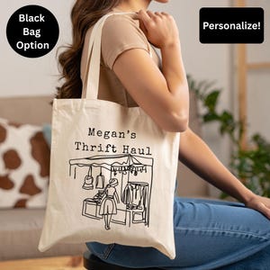 May include: Cream-colored tote bag with the text "Megan's Thrift Haul" and a black line drawing of a thrift store scene. The bag has long handles and the option to personalize it. A black bag option is also available.
