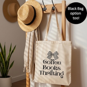 May include: A beige tote bag with a black bow graphic and the words "Coffee Books Thrifting" printed on it. The bag hangs on a wooden rack with a straw hat and scarf. A black circle with the text "Black Bag option too!" is in the upper right.