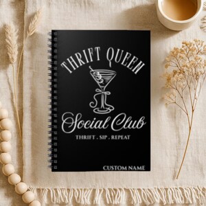 K&ouml;nnte beinhalten: Schwarzes Spiralnotizbuch mit dem Text "THRIFT QUEEN Social Club" in wei&szlig;er Schrift. Das Notizbuch zeigt eine Martini-Glas-Grafik und den Satz "THRIFT. SIP. REPEAT." Auf der Unterseite des Notizbuchs stehen die Worte "CUSTOM NAME".