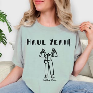 May include: A light green t-shirt with the text "HAUL YEAH!" in black block letters. Below the text is a black and white illustration of a person holding shopping bags. The words "Thrifting Queen" are printed below the illustration.