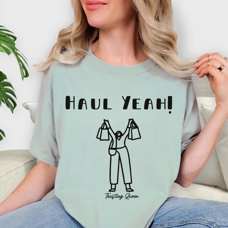 May include: A light green t-shirt with the text "HAUL YEAH!" in black block letters. Below the text is a black and white illustration of a person holding shopping bags. The words "Thrifting Queen" are printed below the illustration.