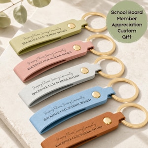 May include: A collection of keychains in various colors: light green, pink, white, light blue, and brown. Each keychain has a gold-colored ring and a leather strap with the text "Shaping Futures, Serving Community ROCKDALE I.S.D. SCHOOL BOARD".