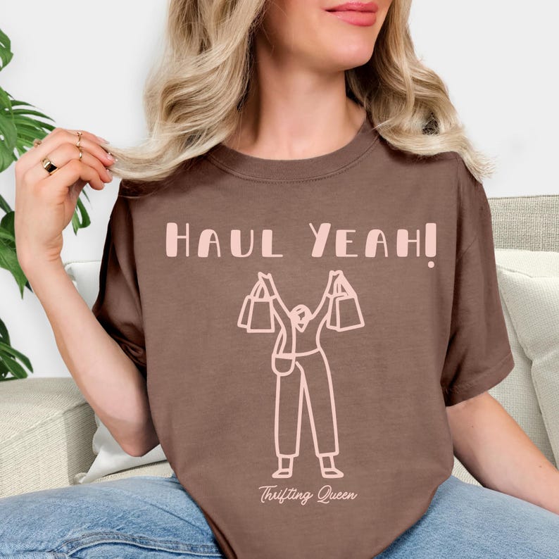 May include: A brown t-shirt with the text "HAUL YEAH!" in light pink. Below the text is a light pink graphic of a person holding shopping bags. The words "Thrifting Queen" are printed below the graphic.