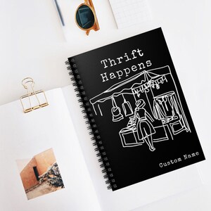 K&ouml;nnte beinhalten: Ein schwarzes Spiralnotizbuch mit den wei&szlig;en Worten "Thrift Happens". Das Cover zeigt eine wei&szlig;e Strichzeichnung einer Marktszene mit einer Person, die einkauft. Die Worte "Custom Name" stehen unten.
